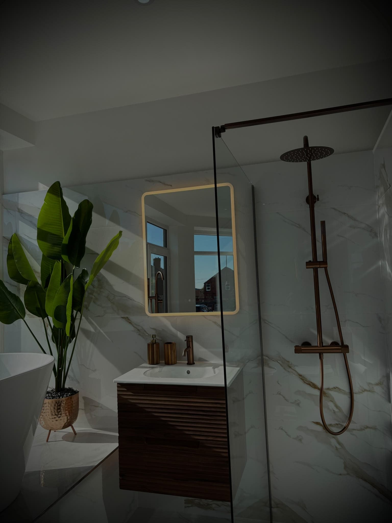 Fluted walnut wall-hung vanity with LED mirror beside a copper walk-in shower